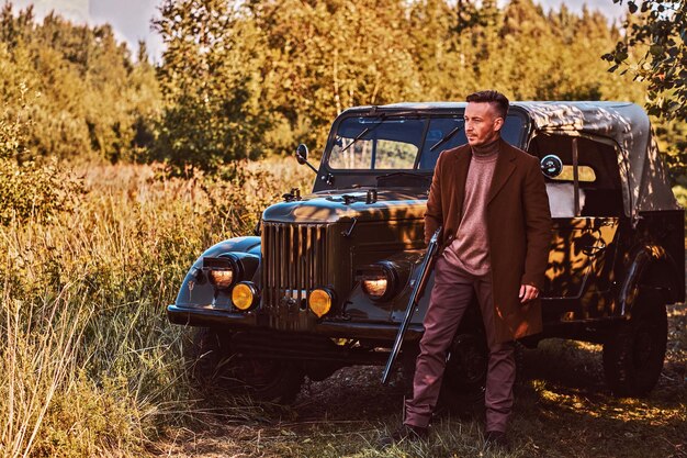 Old fashioned hunter standing together with his beagle dog near a retro military car in a forest.