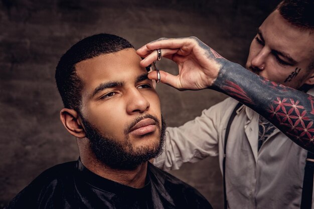 Old-fashioned professional tattooed hairdresser does a haircut to an African American client, using scissors and comb. Isolated on dark textured background.