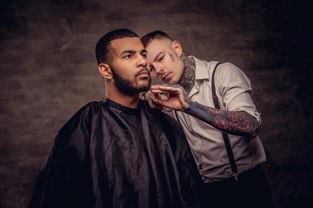 Old-fashioned professional tattooed hairdresser does a haircut to an African American client, using scissors and comb. Isolated on dark textured background.