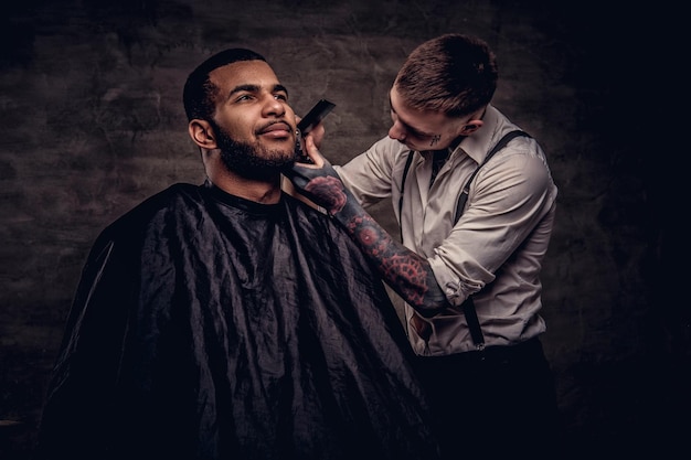Old-fashioned professional tattooed hairdresser does a haircut to an African American client, using a trimmer and comb. Isolated on dark textured background.