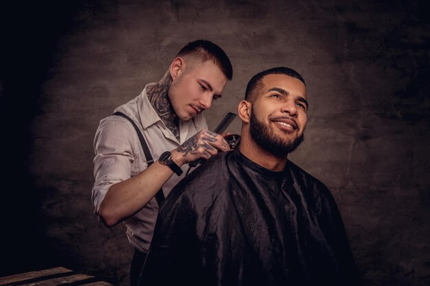 Old-fashioned professional tattooed hairdresser does a haircut to an African American client, using a trimmer and comb. Isolated on dark textured background.