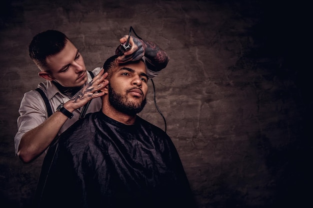 Old-fashioned professional tattooed hairdresser does a haircut to an African American client, using a trimmer and comb. Isolated on dark textured background.