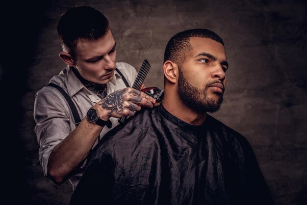 Old-fashioned professional tattooed hairdresser does a haircut to an African American client, using a trimmer and comb. Isolated on dark textured background.