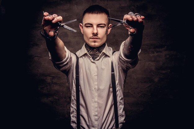 Old-fashioned tattooed hipster wearing a white shirt with suspenders holds a scissors. Isolated on a dark textured background.