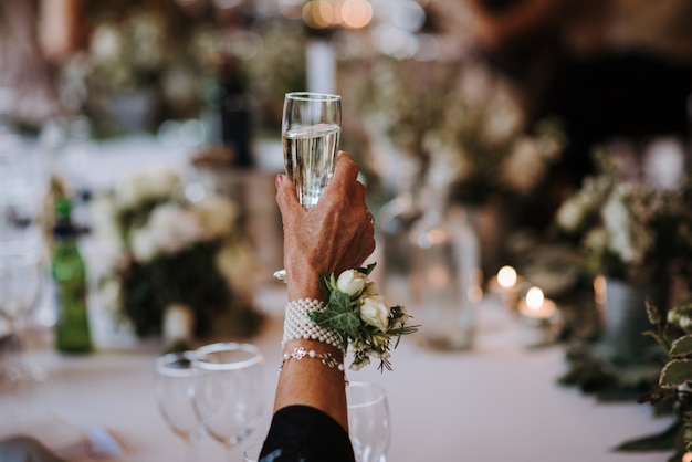 An old female holding a glass of champagne with a flower pinned on an accessory on her hand