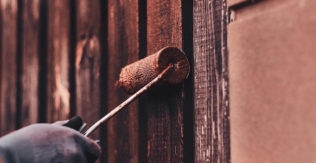 Old fence's renovaiting process by worker. He apply brown paint with brush, wearing protective gloves.