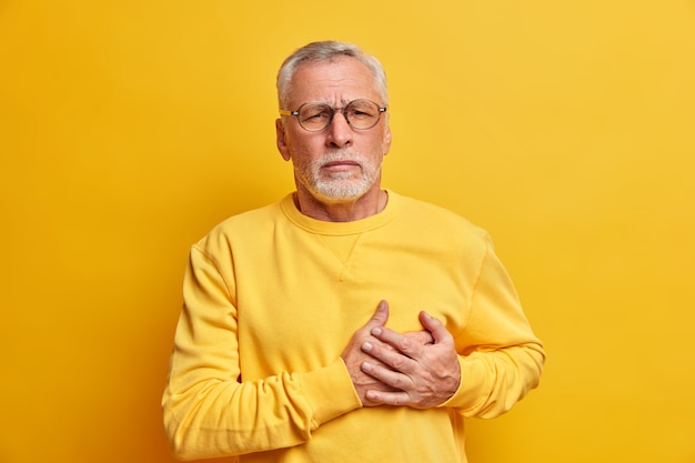 Old grey haired man suffers chest pain has heart attack needs painkillers dressed in casual clothes isolated over vivid yellow wall