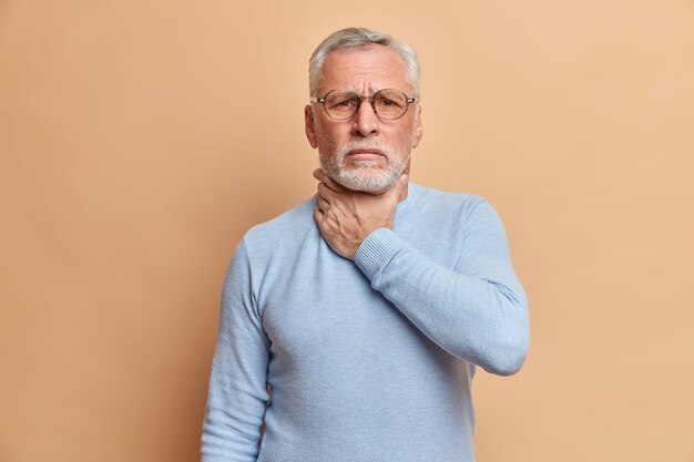 Old handsome bearded man touches neck suffocates because of painful strangle feels pain in throat while swallow wears casual jumper isolated over beige studio wall