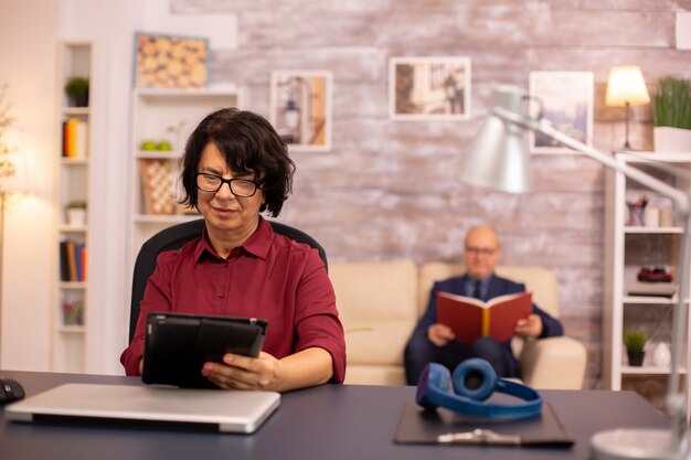 Old lady in her 60s using modern technolgy in cozy living space while her husband reads in the background