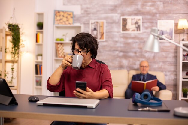 Old lady in her 60s using modern technolgy in cozy living space while her husband reads in the background