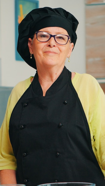 Old lady wearing chef apron and bonete in home kitchen with flour on table for baking looking at camera and smile. Retired elderly baker preparing bakery ingredients to cook homemade bread and cakes