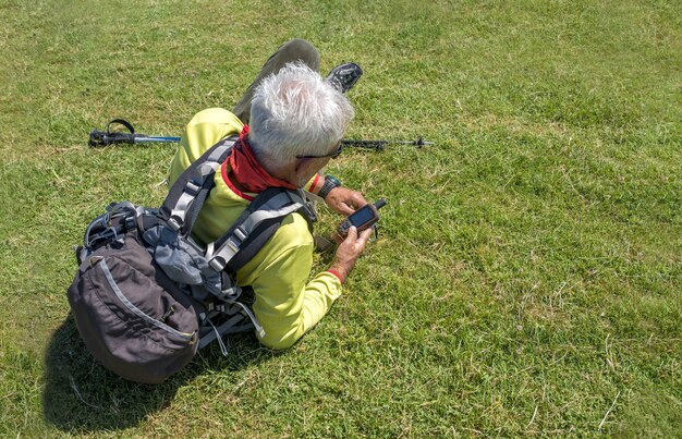 Old male hiker lying on a meadow and looking at handheld navigator