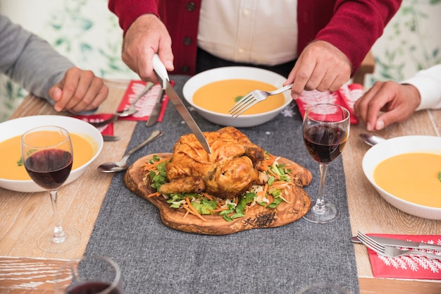 Old man cutting baked chicken on festive table 