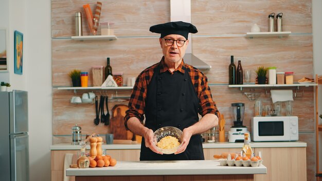 Old man wearing bonete while explaining food preparation recipe looking at camera. Retired blogger chef influencer using internet technology communicating on social media with digital equipment