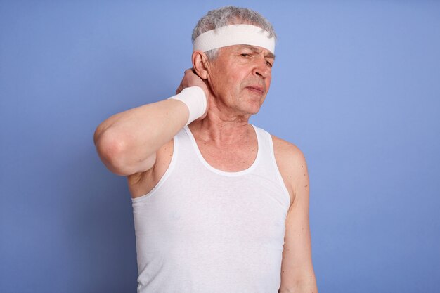 Old man in white t-shirt holding neck in pain isolated, touches her neck, has injury during sport training, sporty male with hairband.