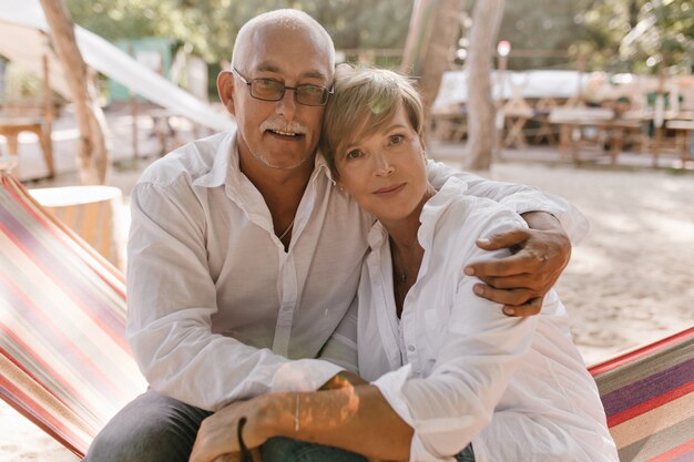 Old man with gray mustache and glasses in shirt looking into camera and hugging blonde short haired woman in white clothes on beach.