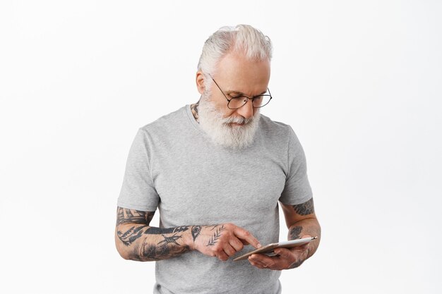 Old man with tattoos reading news, shopping online on gadget, standing against white wall