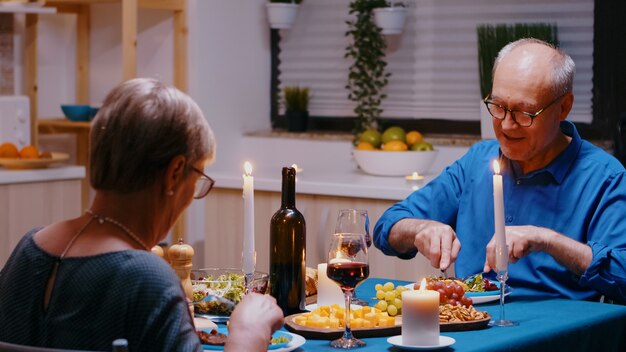 현대 부엌에 있는 테이블에 앉아 낭만적인 저녁 식사를 하는 노부부. 쾌활한 노인들이 이야기하고 식사를 즐기고 식당에서 기념일을 축하합니다.