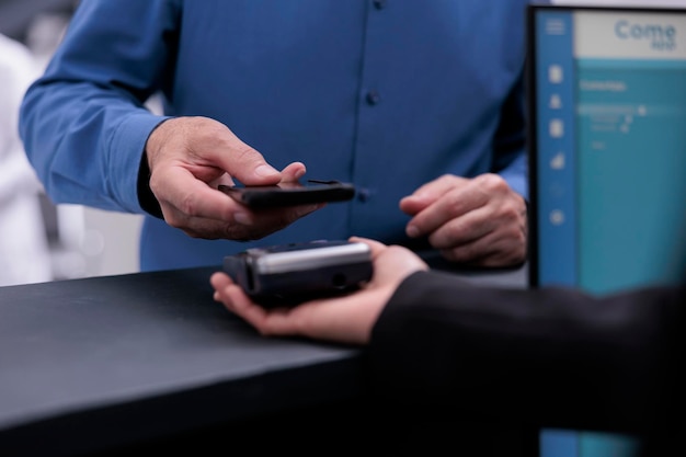 Old patient using mobile phone to pay checkup visit and prescription treatment at hospital reception counter with receptionist. Man paying for examination appointment in waiting area lobby. Close up