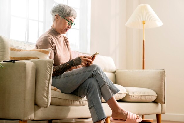 Пожилая пожилая азиатка с татуировкой сидит небрежно, расслабляется, использует смартфон, технологическое устройство, видеозвонок, чтобы поприветствовать и поговорить со своей семьей, белая домашняя изоляция, карантинный период, период