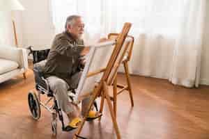 Free photo old senior asian male using art therapy while his legs is injured at homeasian elder man spend recovering of his leg is getting better with painting canvas art in living room at home