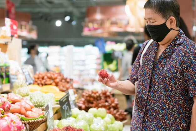 封鎖が終わった後、スーパーマーケットの新しい通常のライフスタイルの食料品店で新鮮なvagetableと果物を買い物している間、古い年配のアジアの女性はウイルス拡散保護マスクを着用します