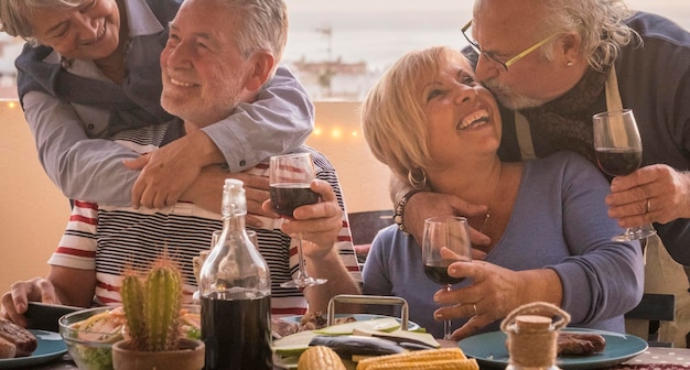食べ物と赤ワインでいっぱいのテーブルの前で一緒に年配の高齢者と友情のライフスタイルのお祝いを愛する幸せな引退した年配の男性と女性は、引退と幸せを楽しんで祝います Premium写真