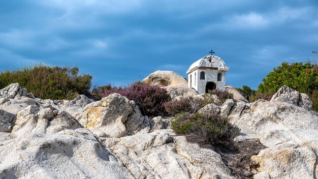 An old and small shrine located on rocks near the Aegean sea coast, bushes around, cloudy sky, Greece