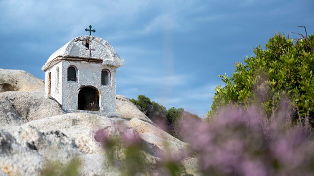 An old and small shrine located on rocks near the Aegean sea coast, bushes around, cloudy sky, Greece