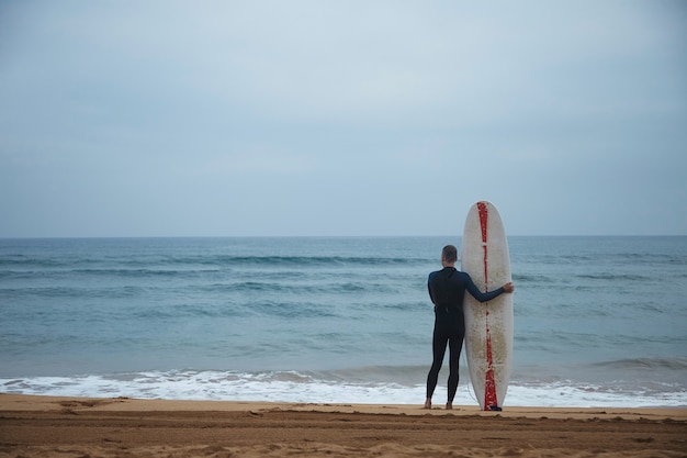 Old surfer with his longboard stays alone on beach in front of ocean and watching waves in ocean before going to surf, wearing full wetsuit in early morning