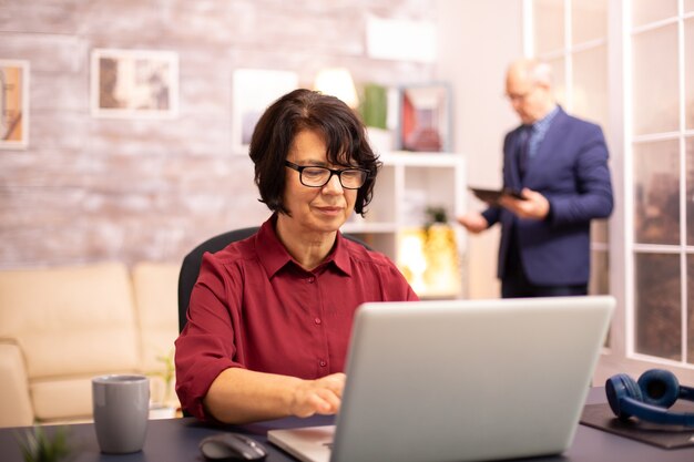 Old woman in her 60s using a modern laptop in her cozy house late in the evening