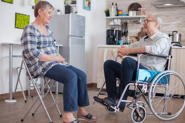 キッチンでおしゃべりをしている車椅子の老婆と障害のある夫。キッチンで夫と会話をしている高齢者。歩行障害のある障害者との生活