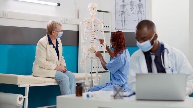 オステオパシーの相談で人間の骨格の骨を分析するフェイスマスクを付けた老婆と看護師。健康診断で解剖学的脊髄の整形外科的診断を説明する医療従事者。