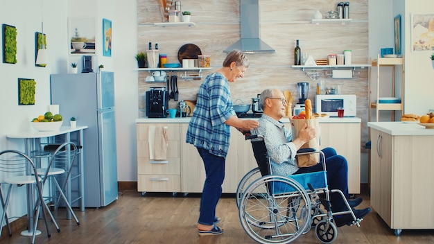 Old woman pushing invalid senior husband in wheelchair after arriving with grocery paper bag from supermarket. Mature people with fresh vegetables for cooking breakfast.