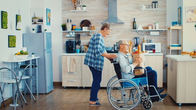 Старая женщина толкает старшего мужа-инвалида в инвалидной коляске после прибытия с продуктовым бумажным пакетом из супермаркета. Зрелые люди со свежими овощами для приготовления завтрака.