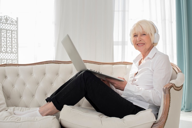 Old woman sitting on sofa with headphones and laptop