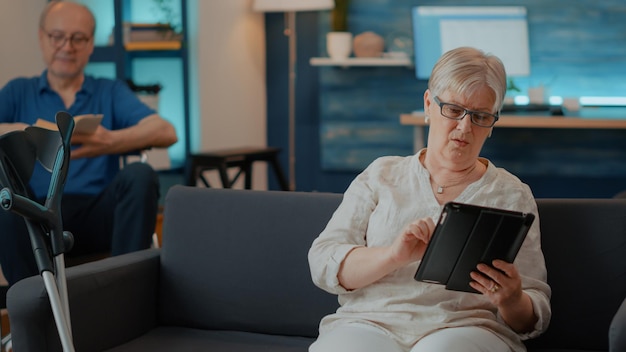 거실에서 터치 스크린이 있는 디지털 태블릿을 사용하는 노부인. 소파에 목발을 짚고 장치로 인터넷을 검색하는 노인은 집에서 재미를 느끼고 은퇴를 즐깁니다. 현대인