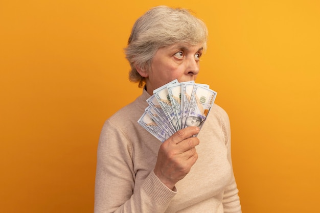 Пожилая женщина в сливочном свитере с высоким воротом стоит в профиль и держит деньги, глядя сзади, изолированную на оранжевой стене с копией пространства