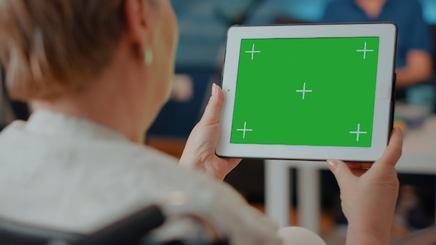 Old woman with disability holding green screen on digital tablet. Senior person sitting in wheelchair, using blank copy space with isolated chroma key and mock up background on display.