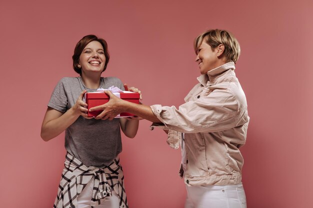 Old woman with short hair in beige jacket gives red gift box to joyful girl in grey tshirt and plaid shirt on pink background