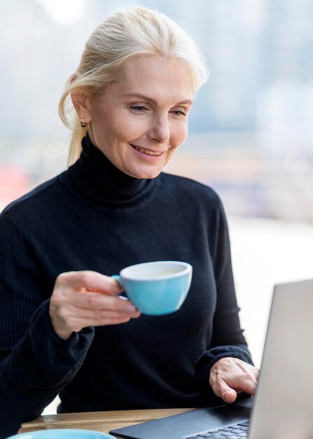 Older business woman enjoying coffee outdoors while working on laptop