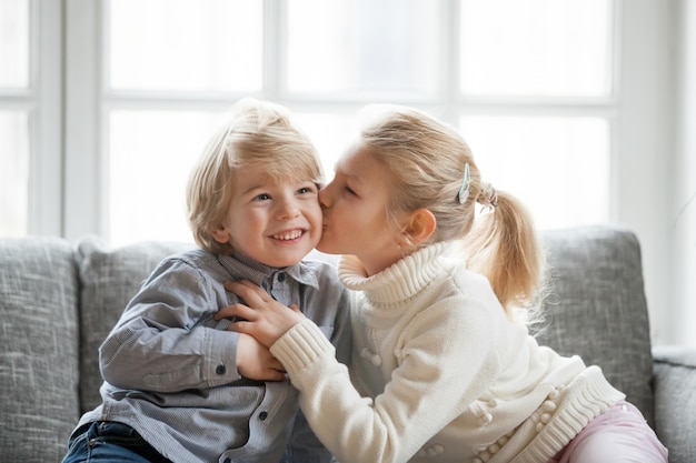 Older child sister embracing kissing little younger brother at home