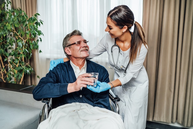 Free photo an older man in a wheelchair smiles at the nurse-assistant, she hands him a glass of water.