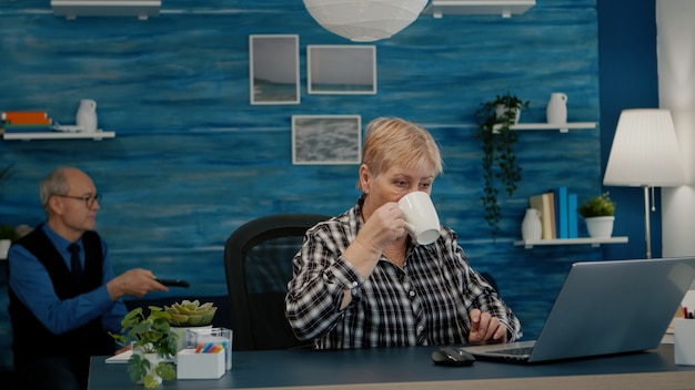 Older professional mature business woman using laptop sitting at workplace desk drinking coffee