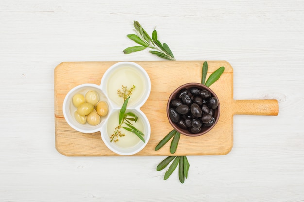 Olives assortment and olive oil with olive leaves in a bowls and cutting board on white wood, top view.