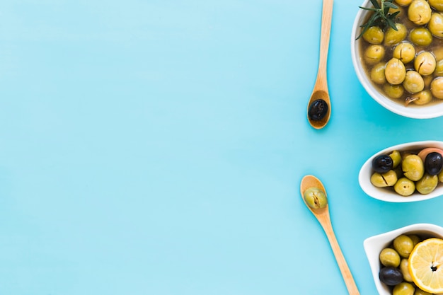 Olives in different bowls and two wooden spoon over the blue background