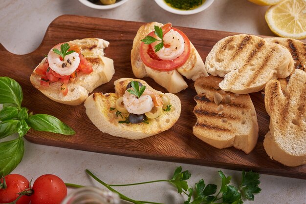 Olives, oil, Grilled shrimps and fresh vegetable salad on a wooden board on black slate stone board over dark surface, juicy tomatoes on fresh bread, pesto as topping. top view. Flat lay