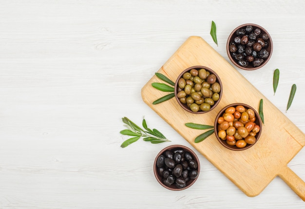 Разнообразие оливок с листьями оливкового дерева в глиняных мисках и разделочной доске на белой древесине, плоская планировка.