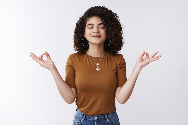 Om girl feels peace patience. Attractive carefree relaxed happy young woman curly shirt hairstyle close eyes smiling delighted meditating hands sideways nirvana lotus pose, breathing practice yoga