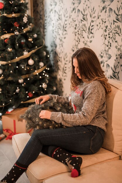 oman in sweater with Christmas wreath near decorated fir tree